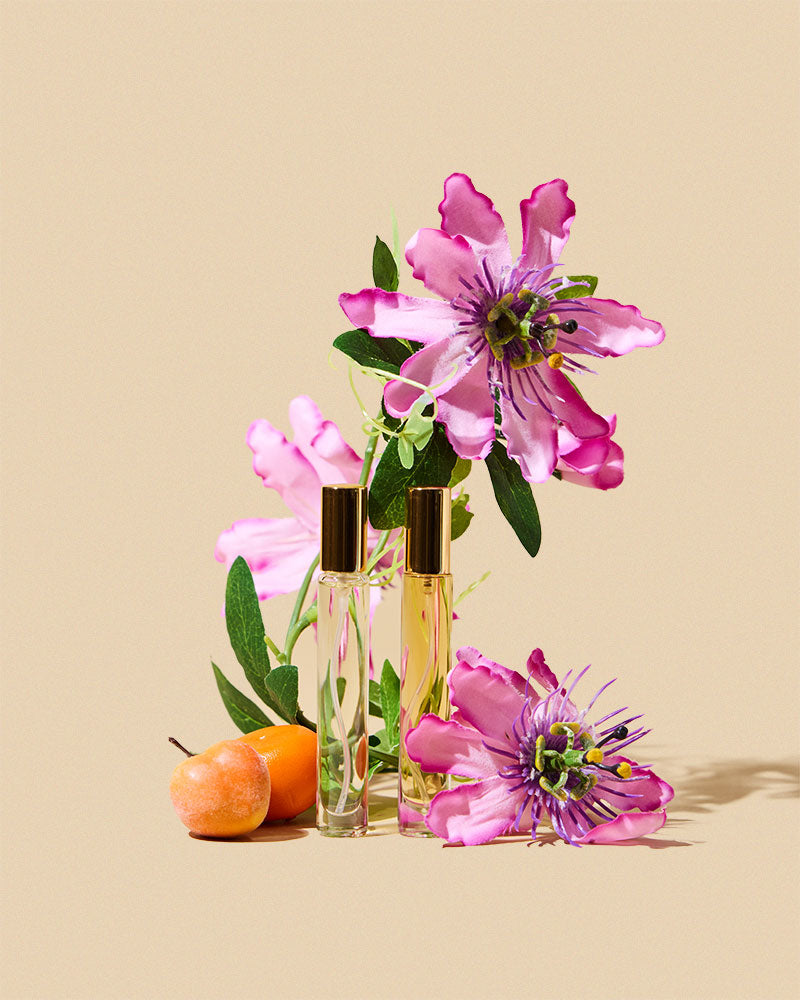 Two small glass bottles with gold caps surrounded by pink flowers on a beige background
