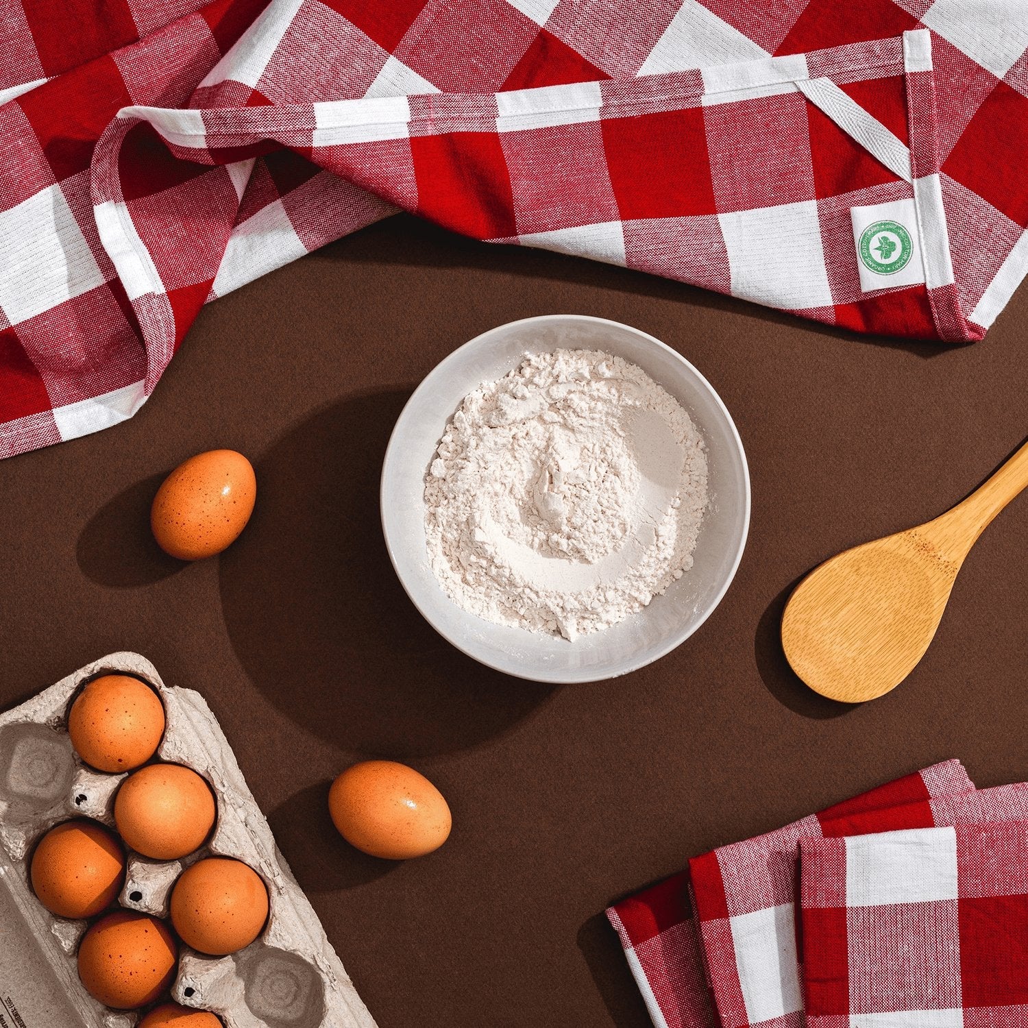 Bowl of flour, eggs, and wooden spoon on a brown surface with red and white checkered cloth.
