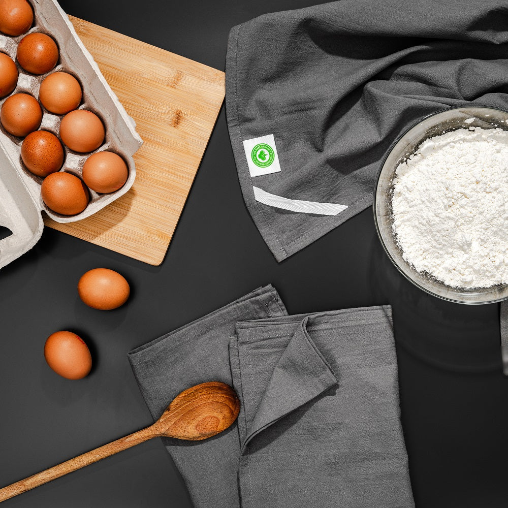 Eggs, a wooden spoon, flour, and gray towels on a dark surface