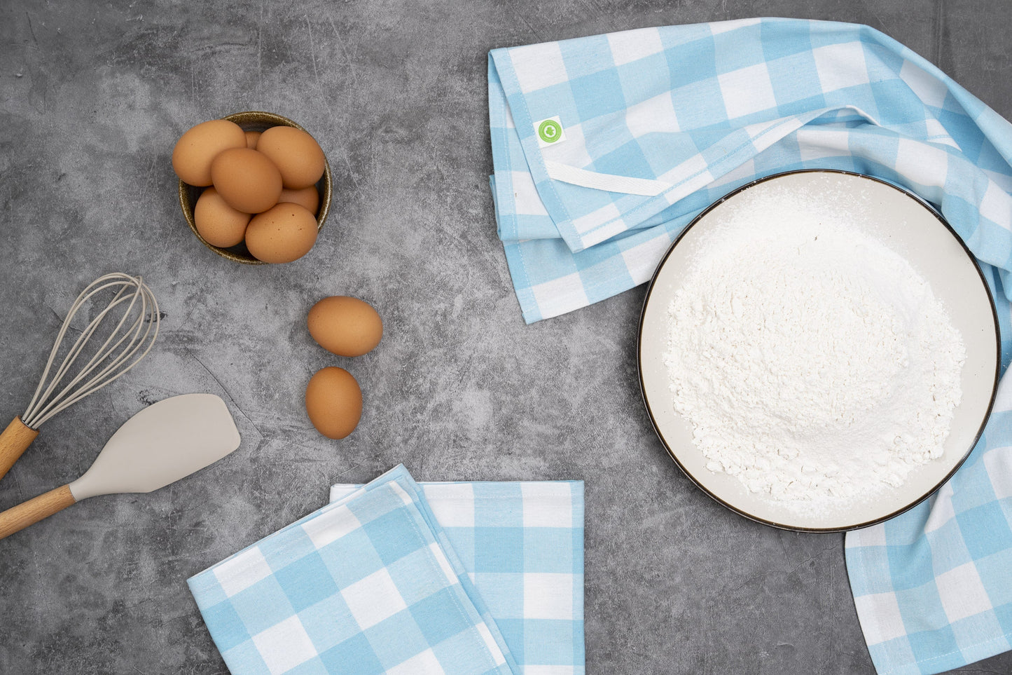 Eggs, a whisk, spatula, and checkered towels on a gray surface
