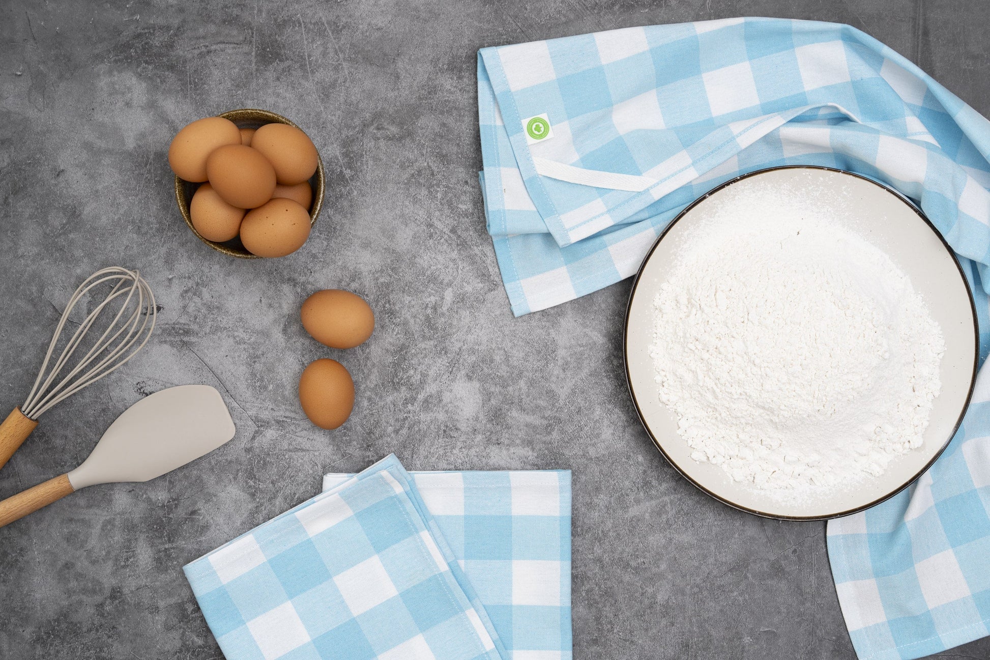 Eggs, a whisk, spatula, and checkered towels on a gray surface