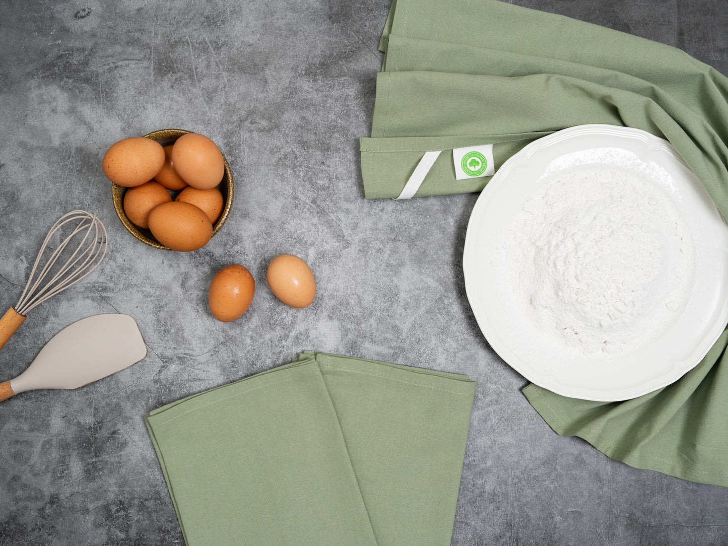 Eggs in a basket with a whisk, spatula, and sage green napkins on a gray surface.