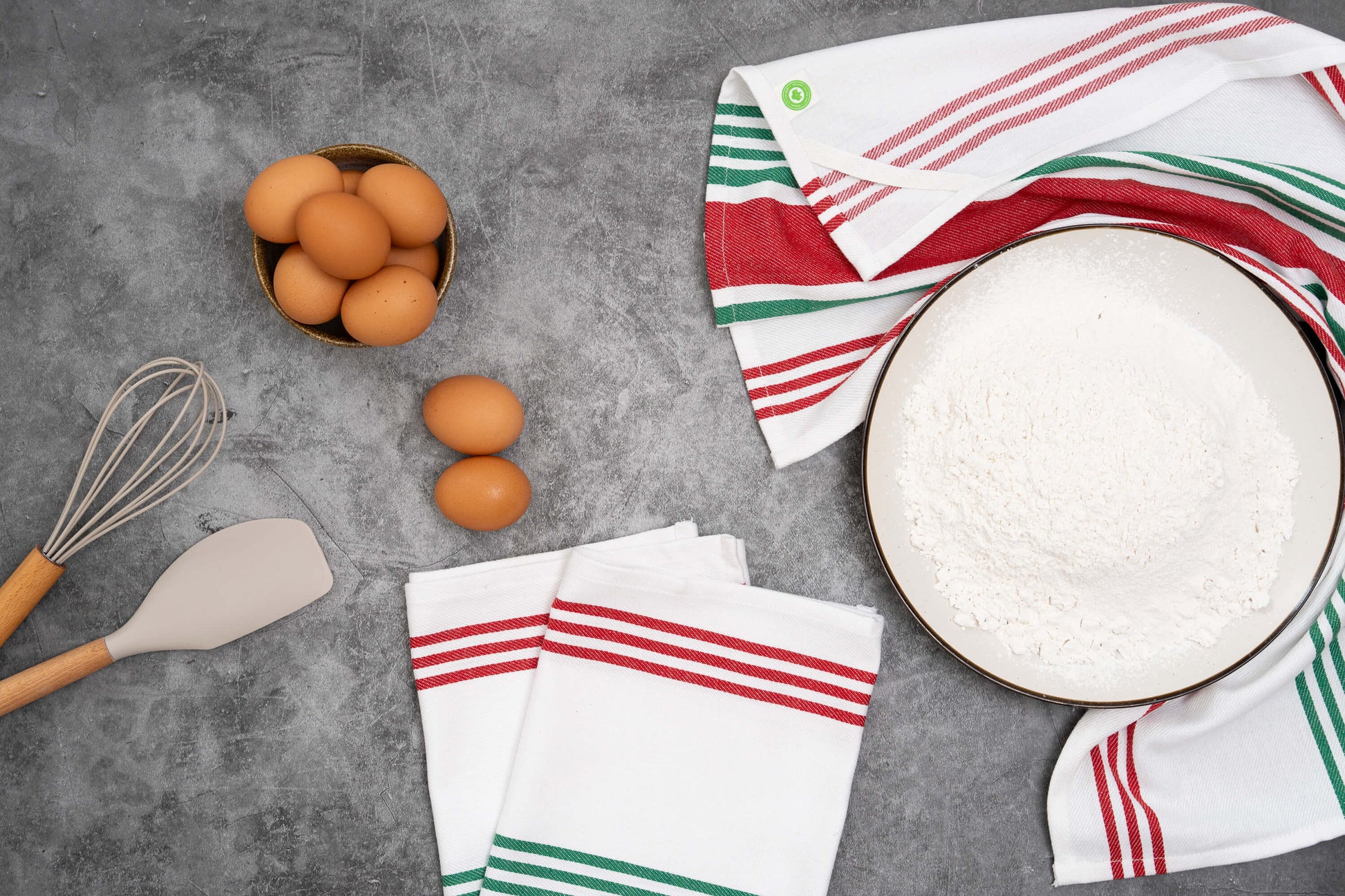 Kitchen items including eggs, a whisk, spatula, and a bowl with a red, white, and green striped towel on a gray surface.