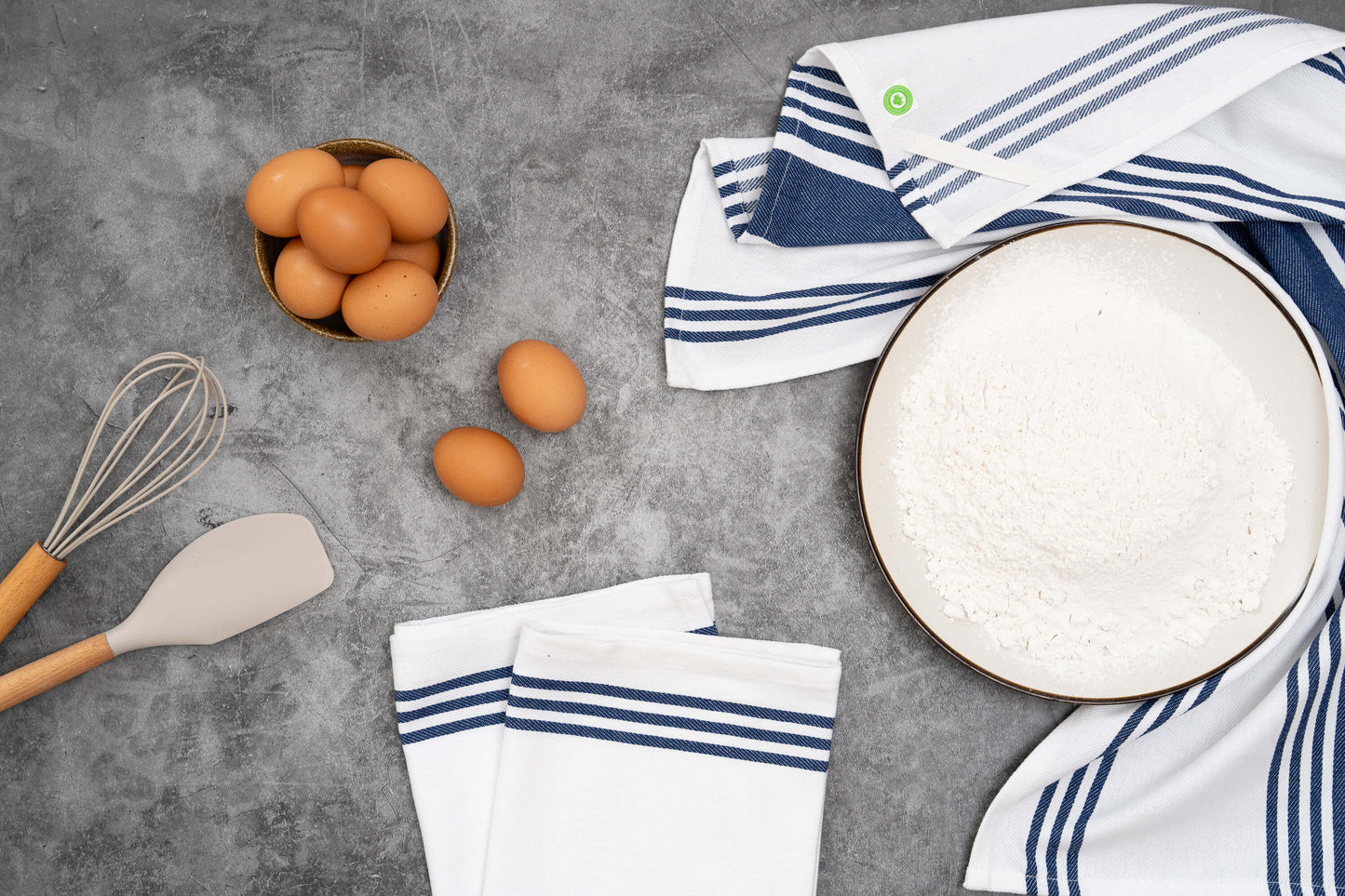 Kitchen items including eggs, a whisk, a spatula, and striped towels on a gray surface.