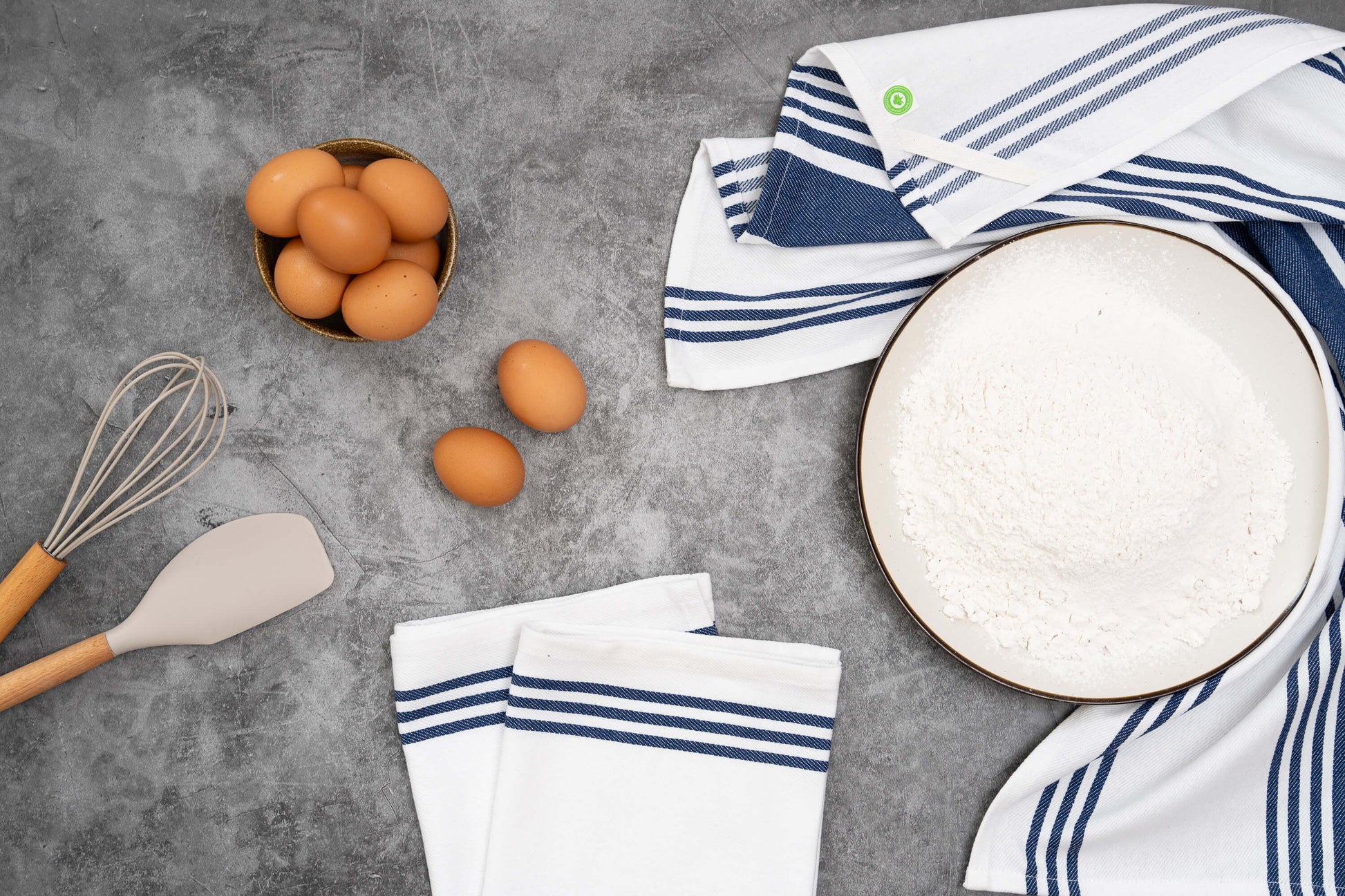 Kitchen items including eggs, a whisk, a spatula, and striped towels on a gray surface.