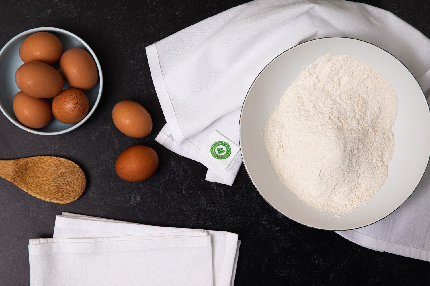 Eggs, flour, and a wooden spoon on a dark surface with a white towel.